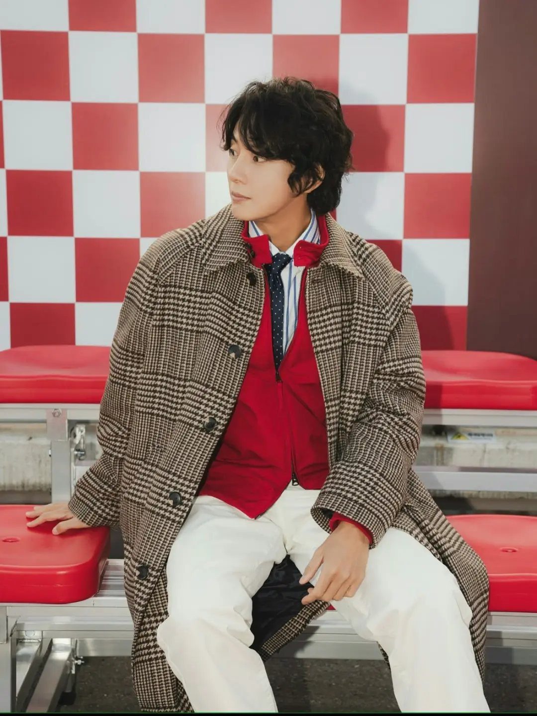 Man wearing a brown plaid trench coat, red sweater, and white pants.
