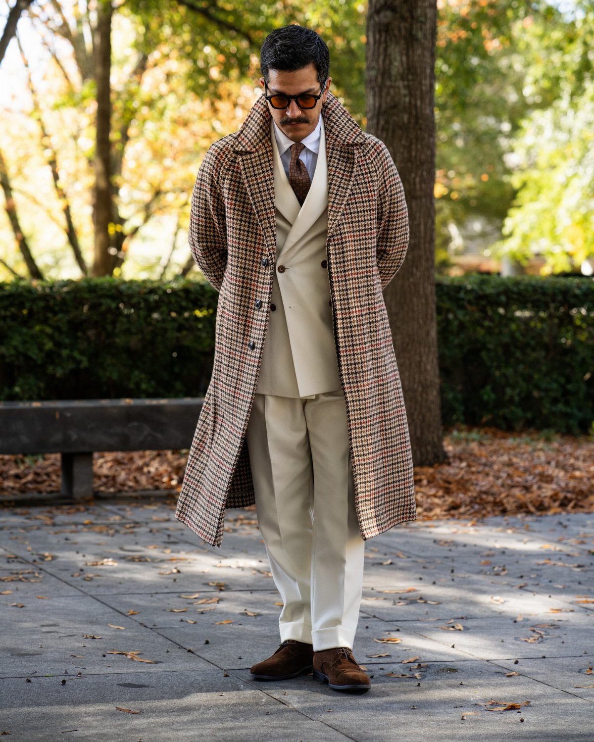 Man in a brown plaid trench coat and cream suit standing outdoors.