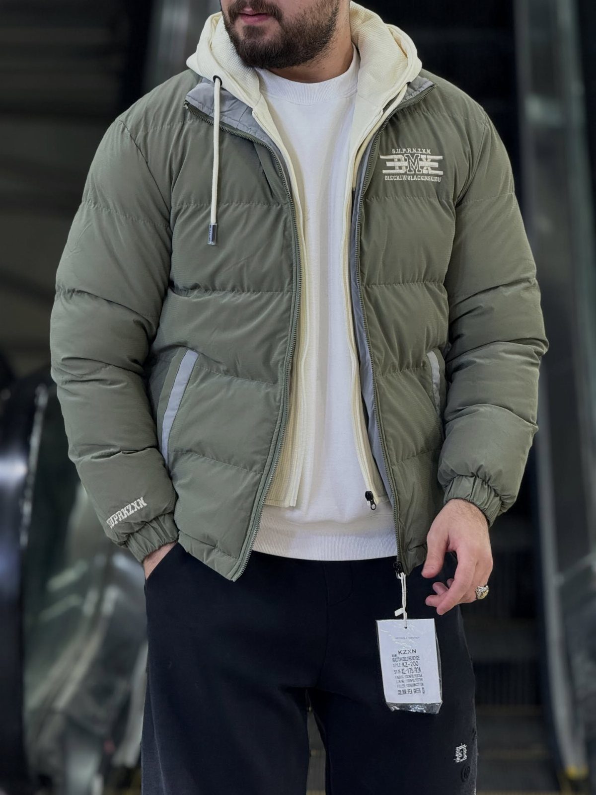 Man wearing olive green puffer jacket over white hoodie and long-sleeve shirt.