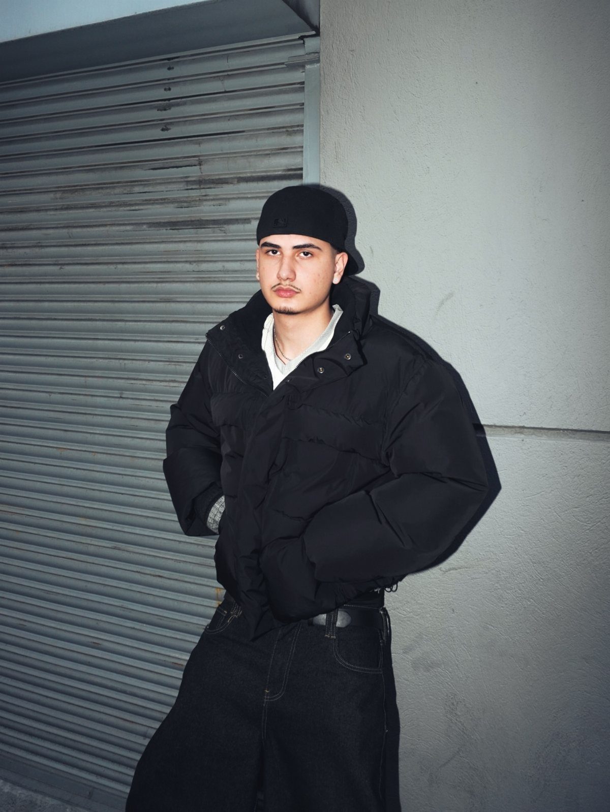 Man in black puffer jacket and black jeans posing against a grey wall.