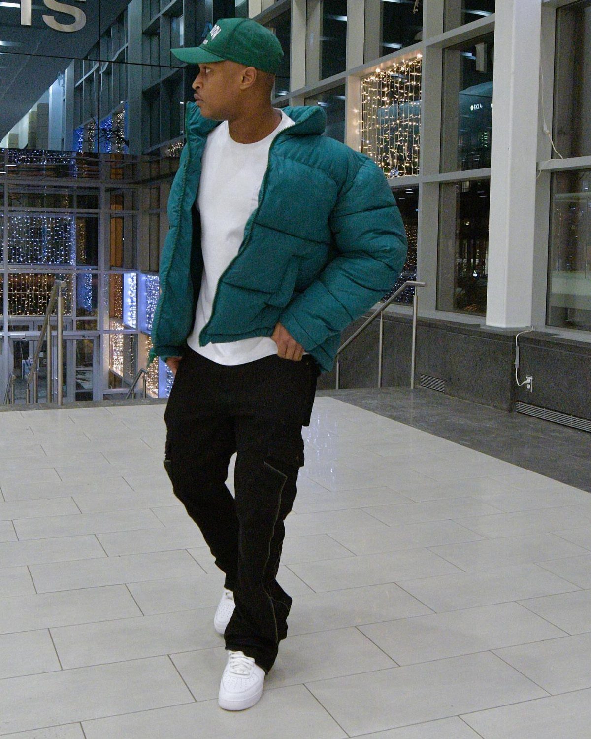Man in teal puffer jacket, black cargo pants, and green baseball cap posing indoors.