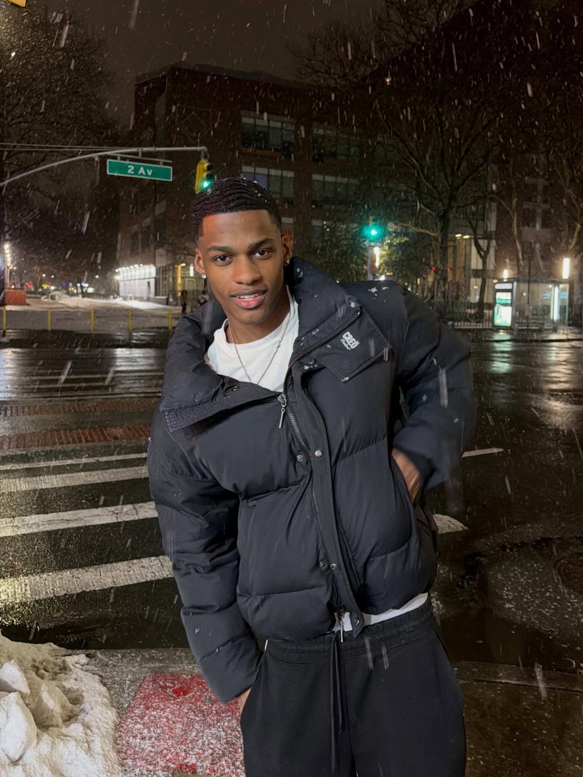 Man in black puffer jacket and black joggers standing in falling snow.