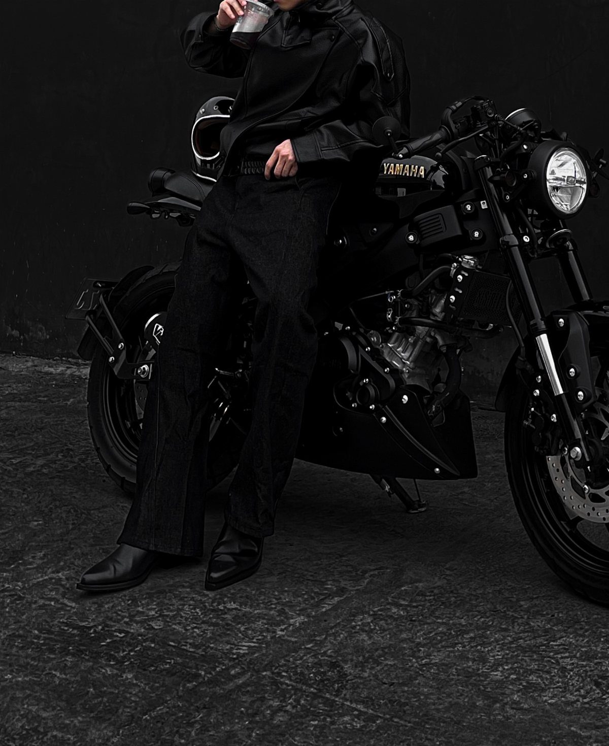 Person in black leather racer jacket and trousers leaning on a black motorcycle.