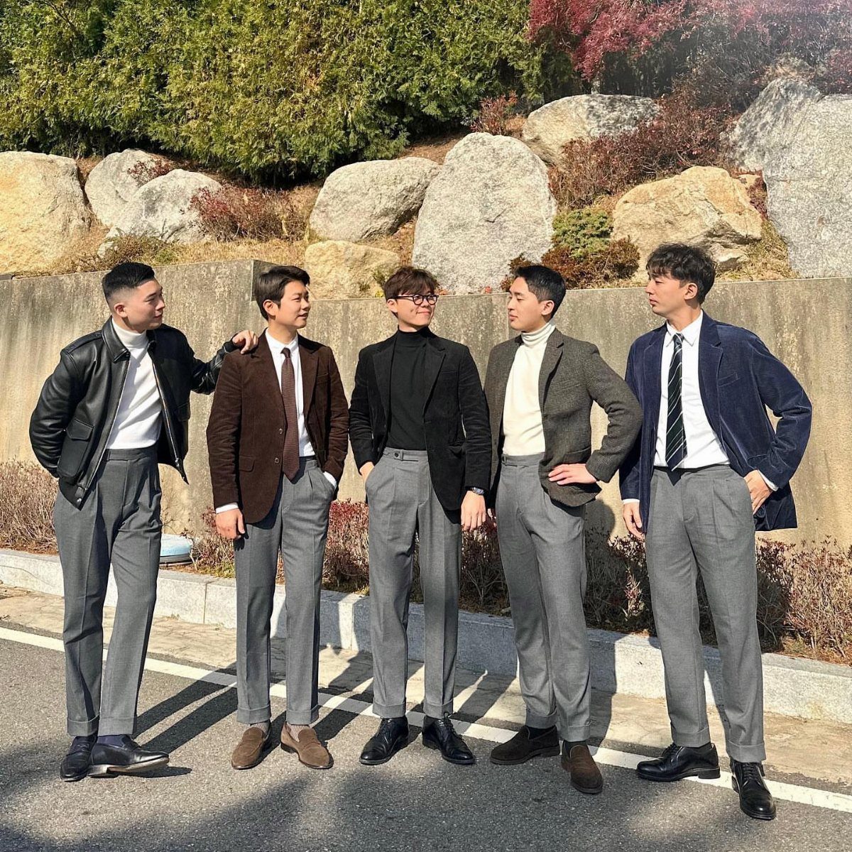 Man in black leather racer jacket and grey dress pants standing with friends.