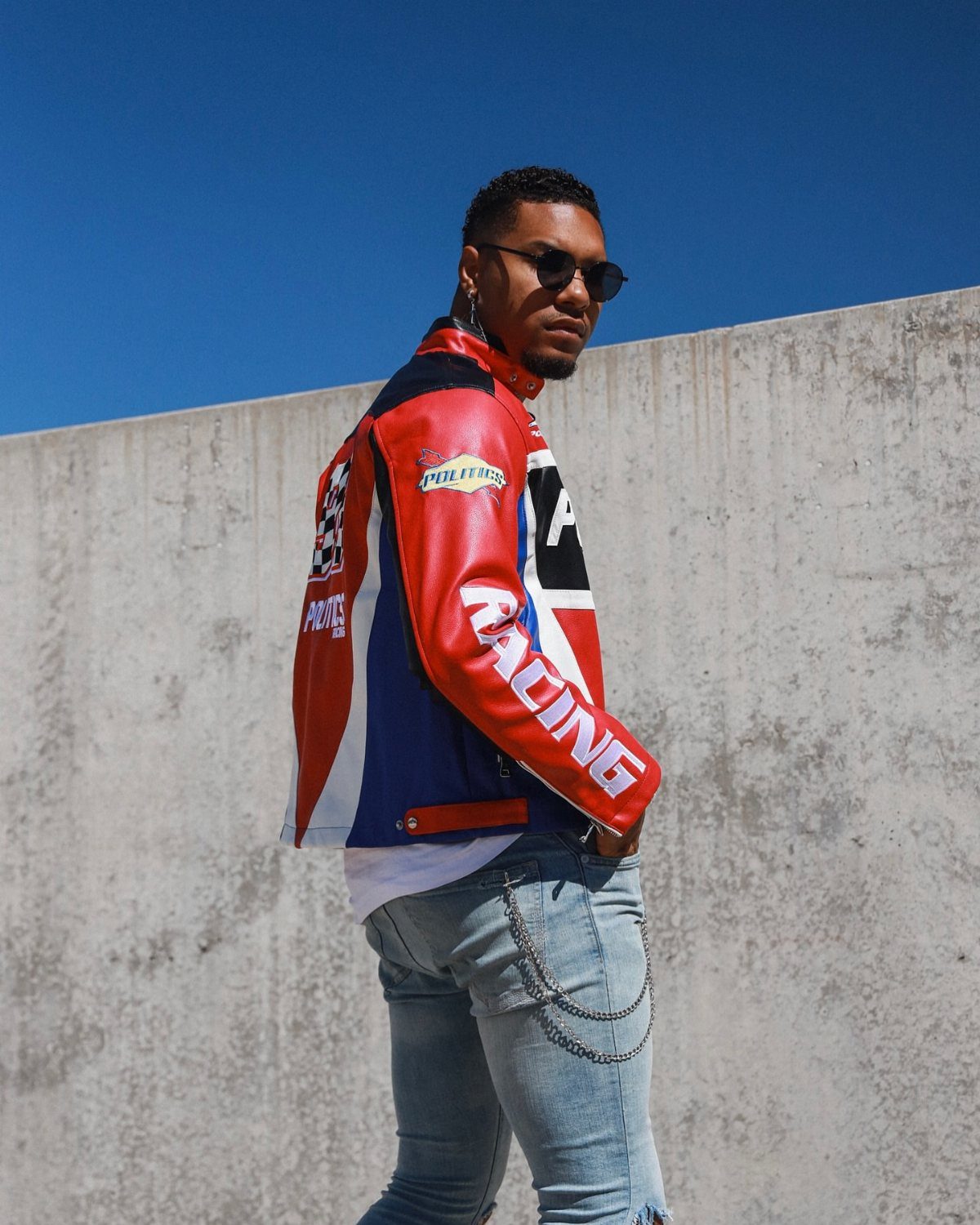 Man in a colorful racing jacket and light blue slim jeans posing outdoors.