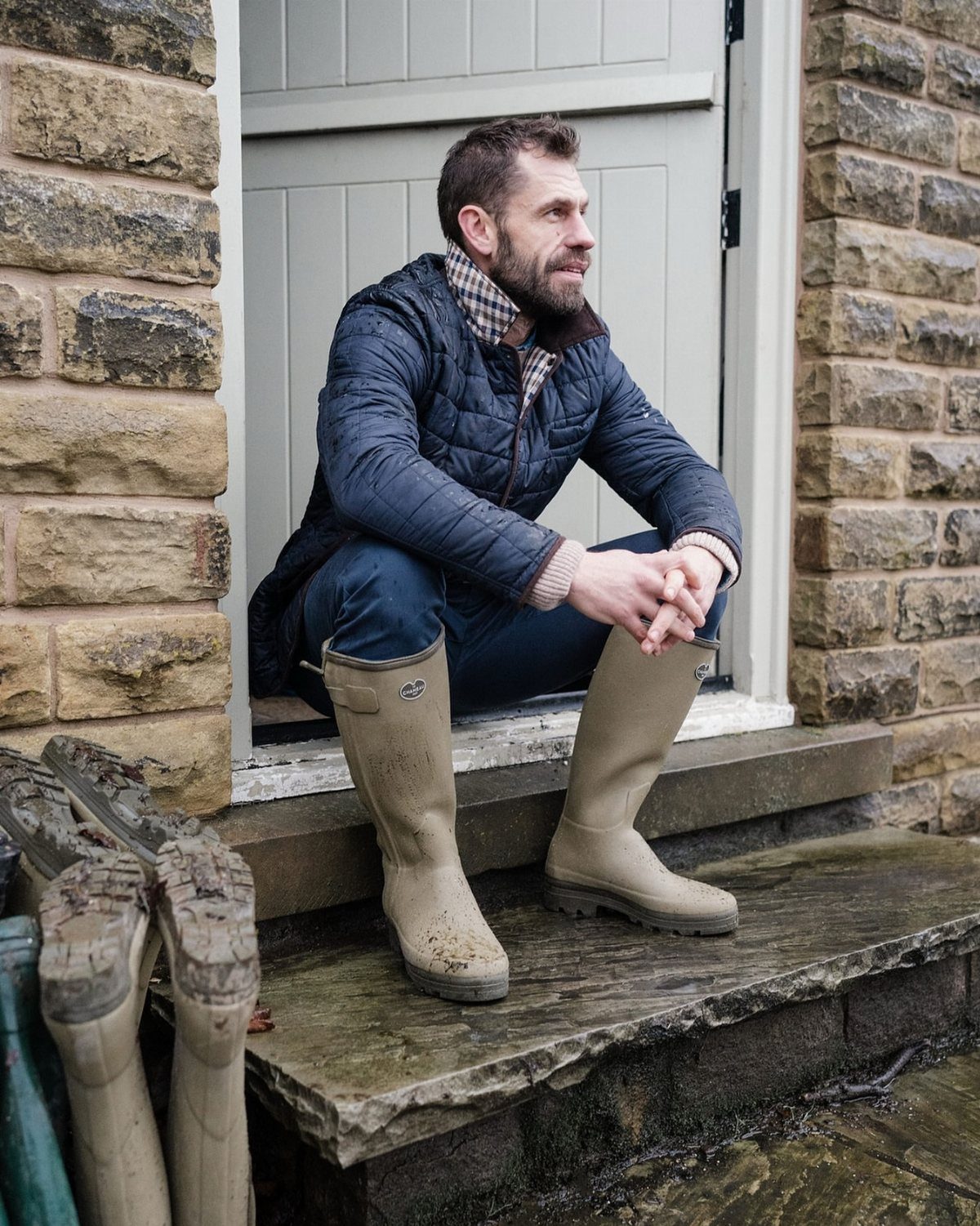 Man sitting on a stone doorstep wearing tall beige rubber rain boots.