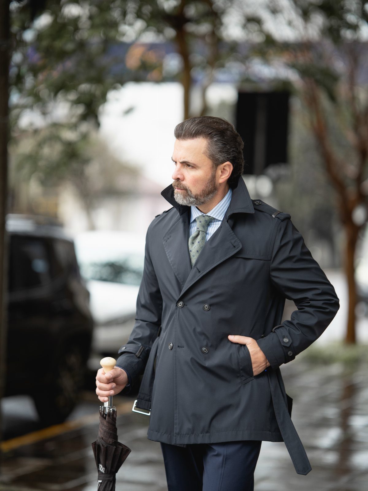 Man in a dark grey trench coat holding a black umbrella on a street