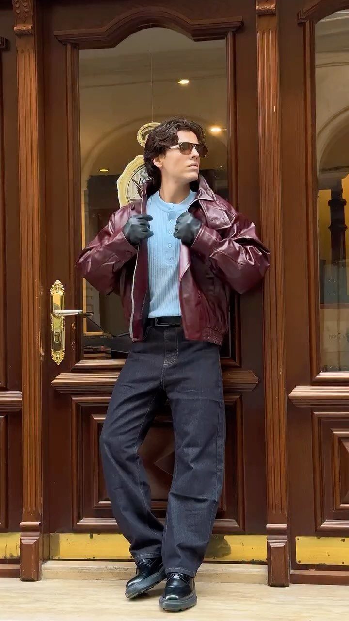 Person wearing a red leather jacket, blue shirt, and black trousers by a door.