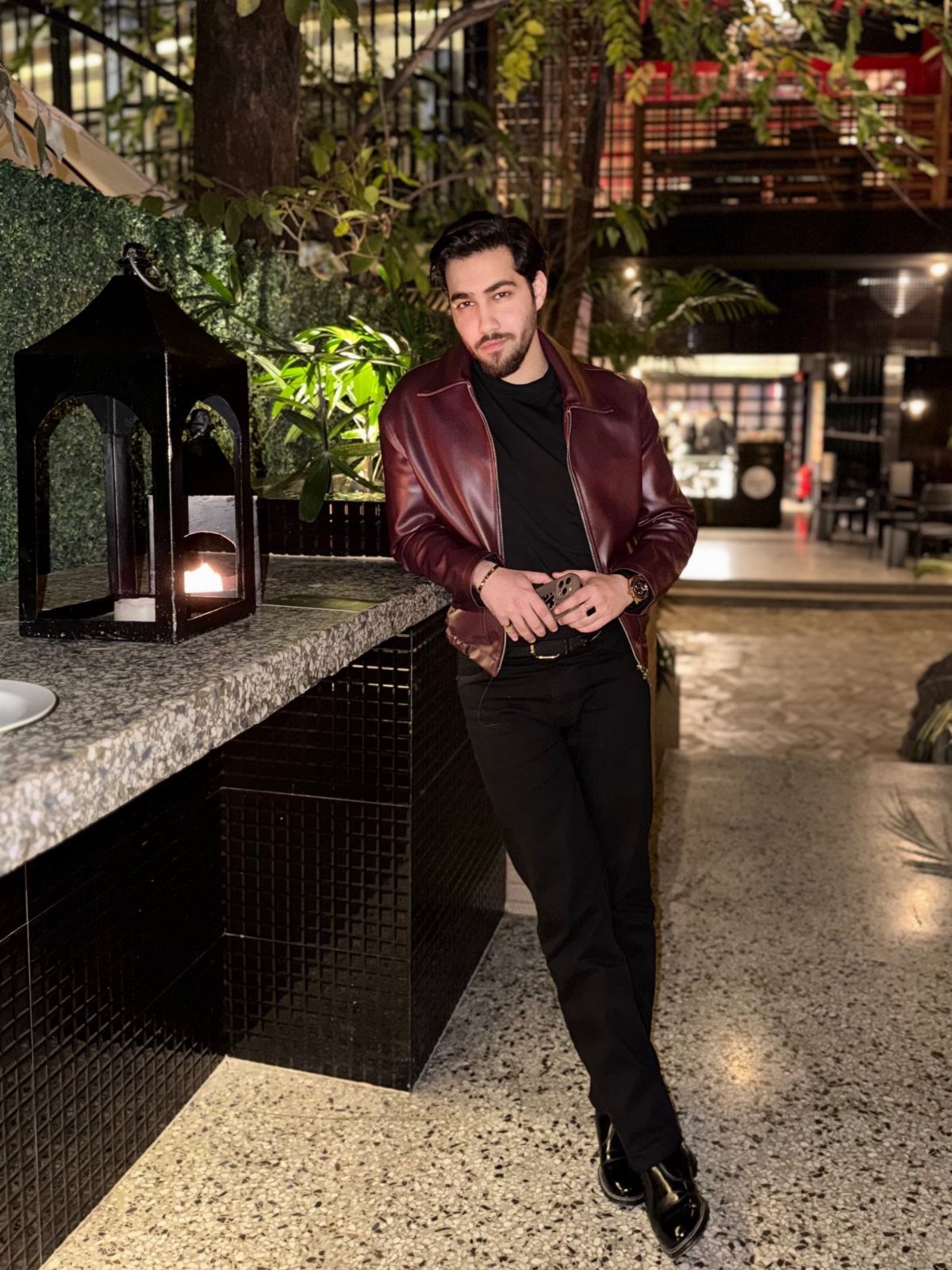 Man in a red leather jacket and black trousers leaning on a counter.