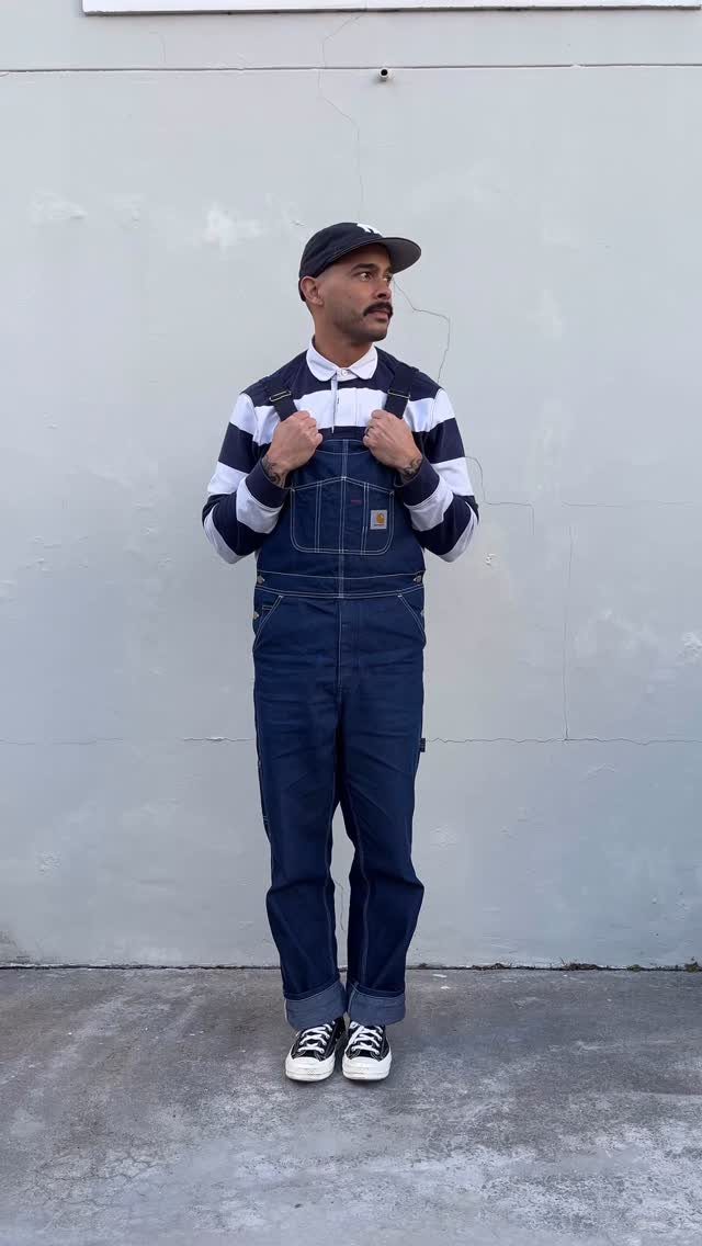 Man wearing dark blue denim overalls and black and white canvas sneakers.