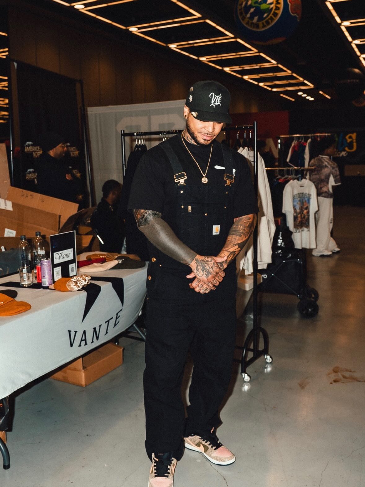 Man wearing black overalls and light-colored sneakers standing in a clothing event space.