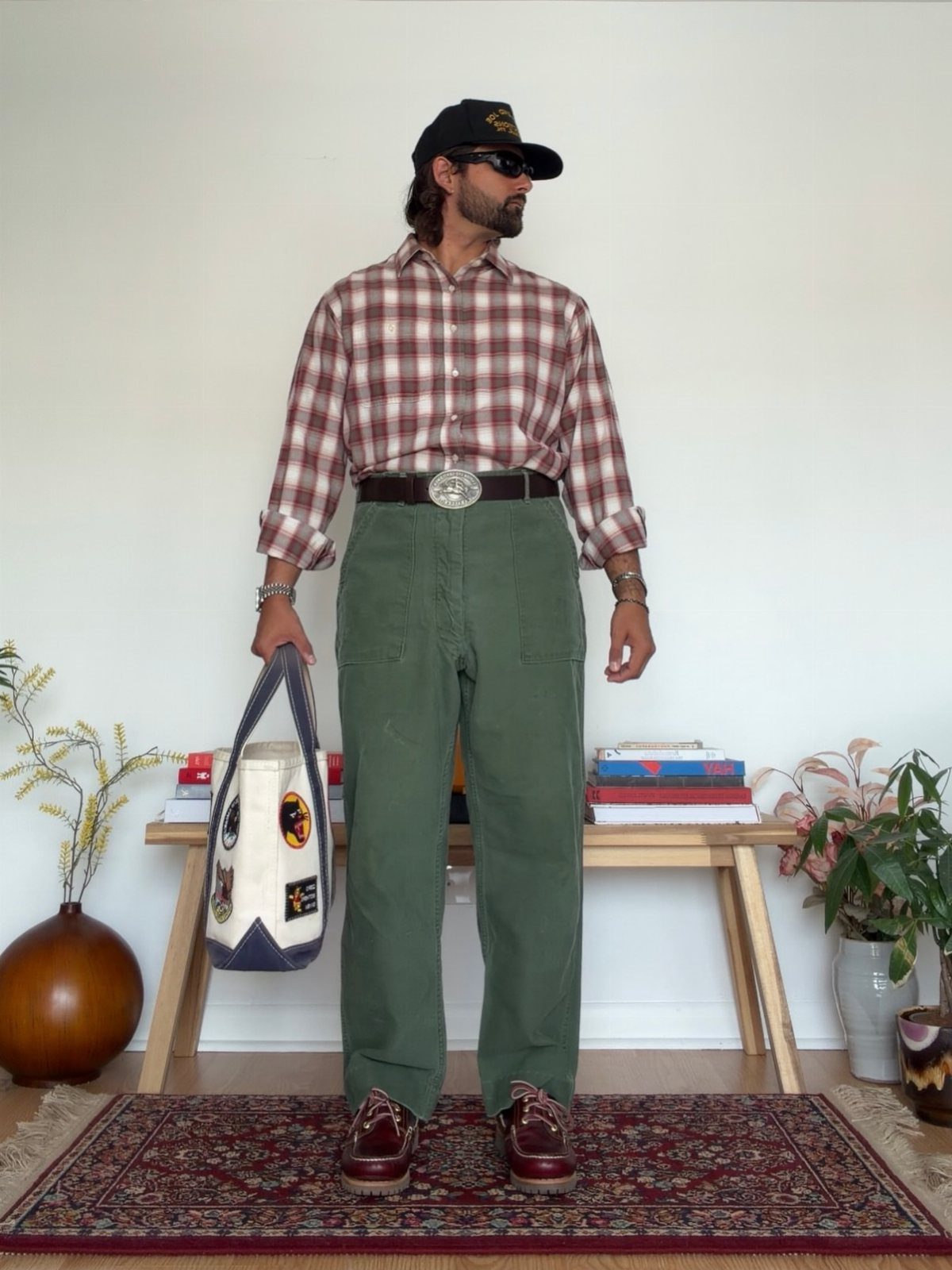 Man in checkered shirt and green trousers holding a tote bag indoors.