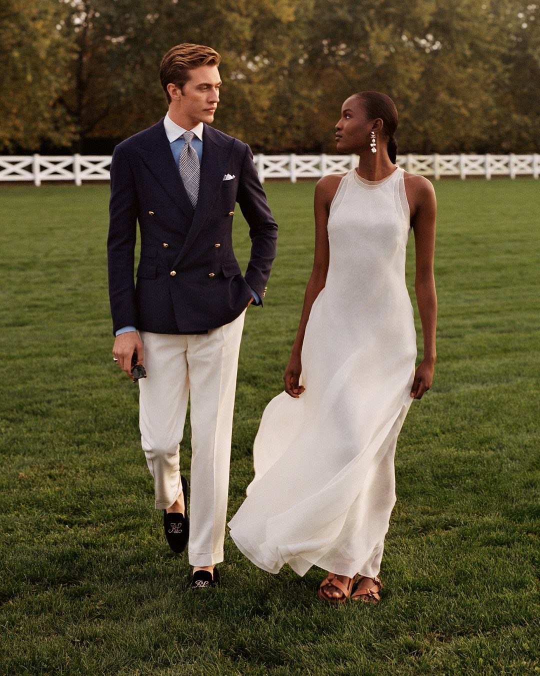 Man in navy blazer and white trousers walking with woman in white dress.