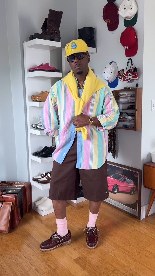 Man in pastel striped shirt, yellow cap, and brown shorts posing indoors.