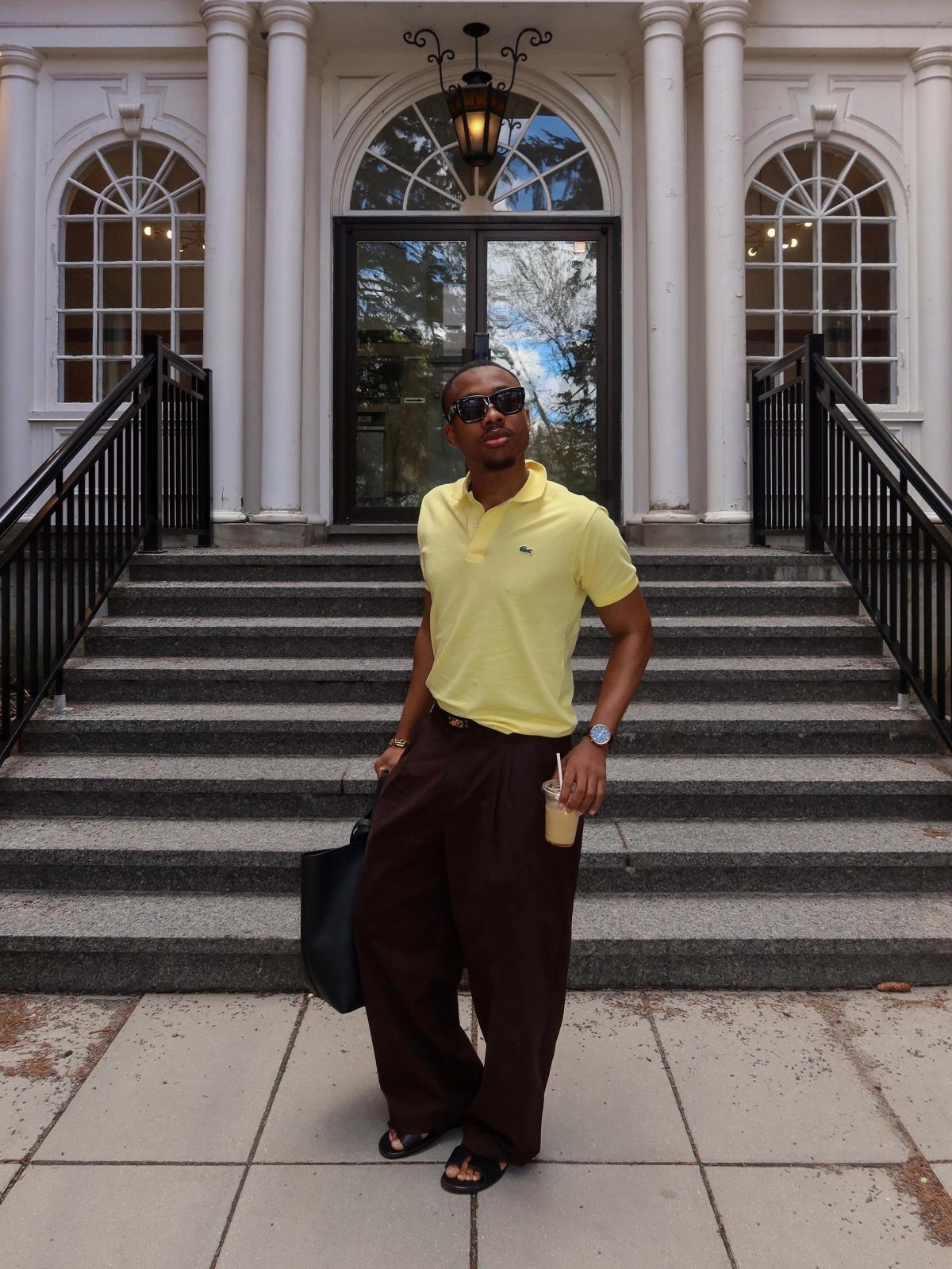 Man in yellow polo and brown wide-leg trousers standing on building steps.