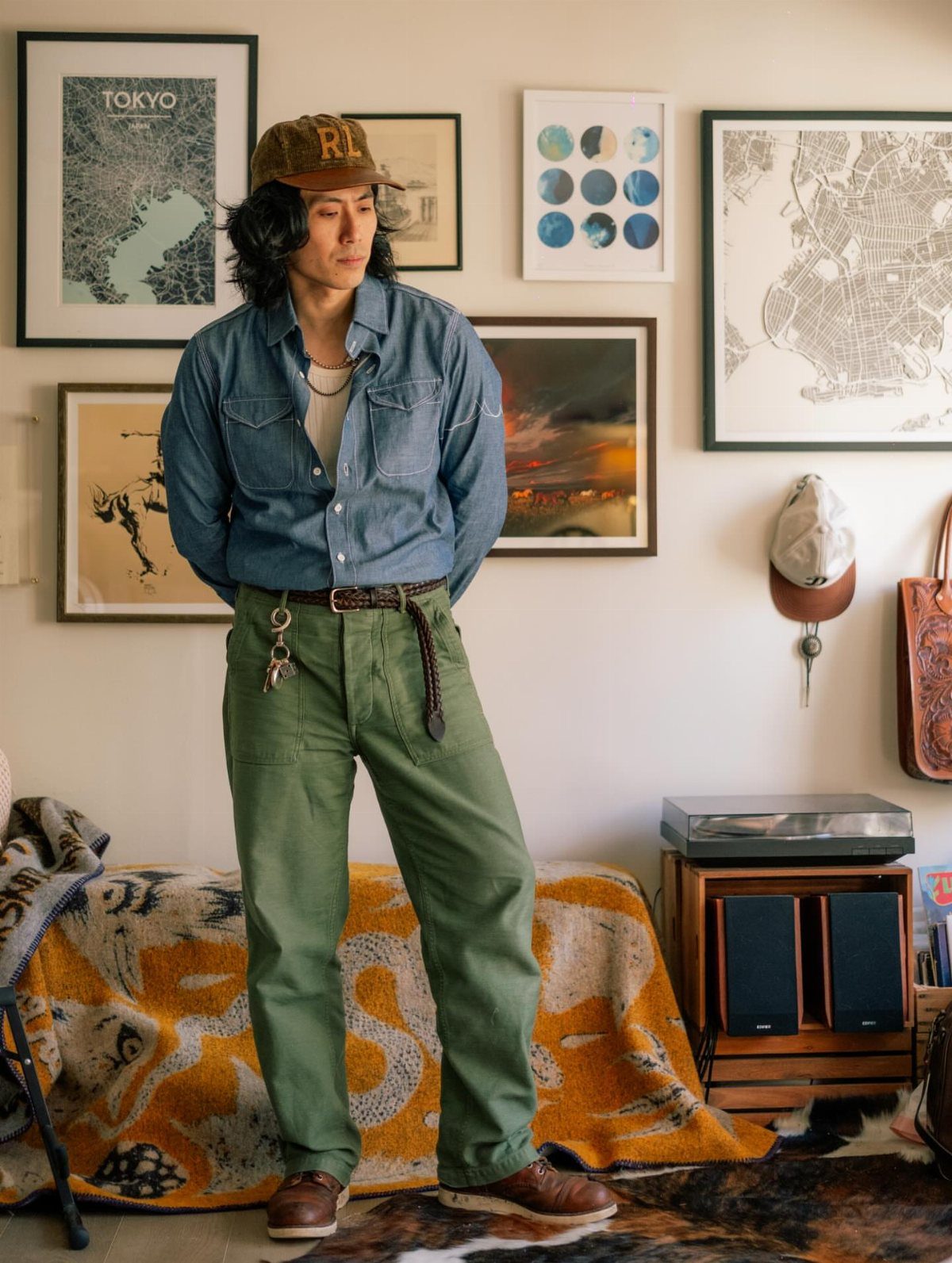 Man wearing a blue denim shirt, olive green pants, and brown cap indoors.