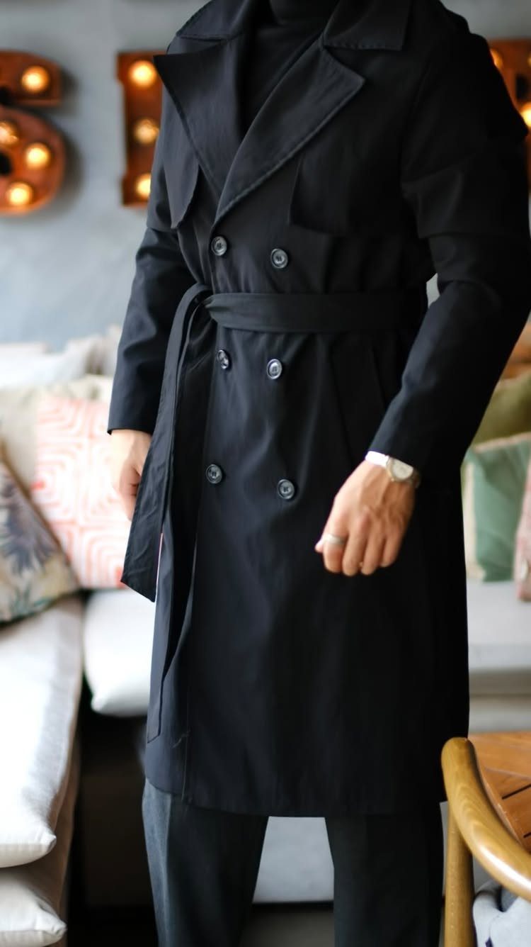 Man wearing a long black belted trench coat and matching black trousers indoors.