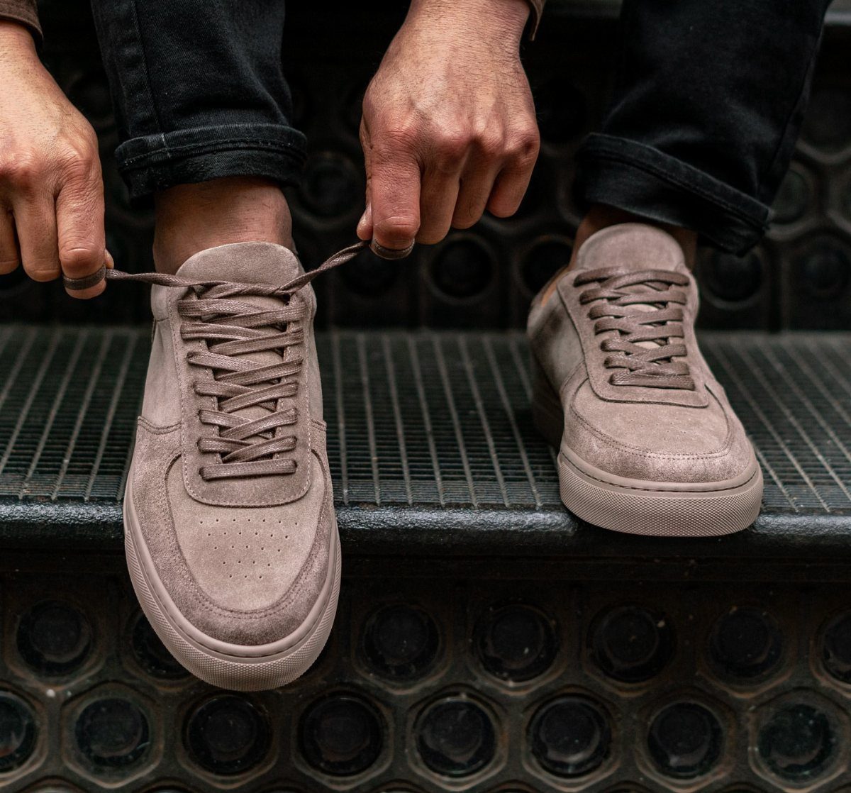 Mauve suede lace-up sneakers worn with black jeans on a metal grate.