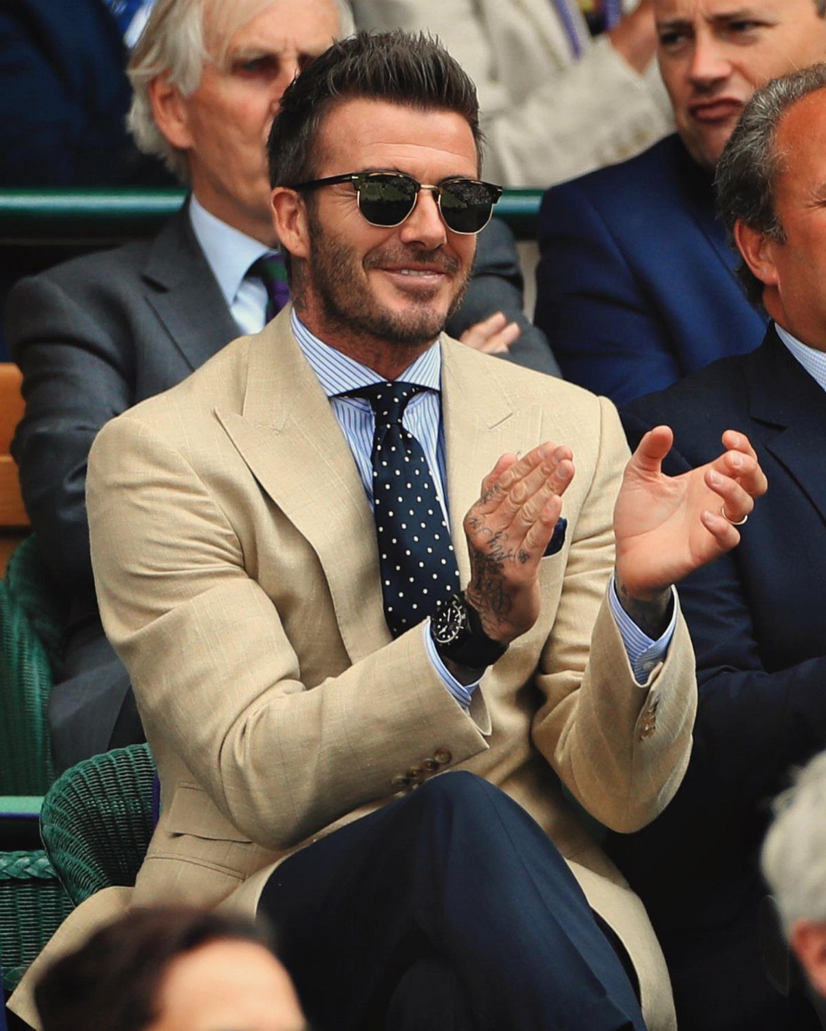 Man in tan linen blazer and polka-dot tie clapping at a sporting event