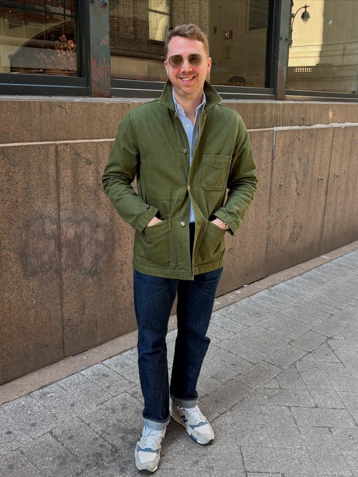 Man wearing olive green jacket, blue shirt, and dark jeans on city sidewalk