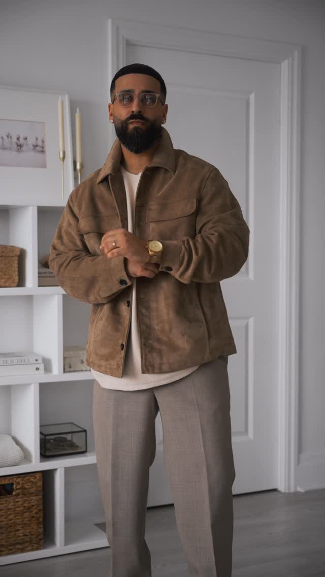 Man wearing a brown suede overshirt and grey trousers in a white room.