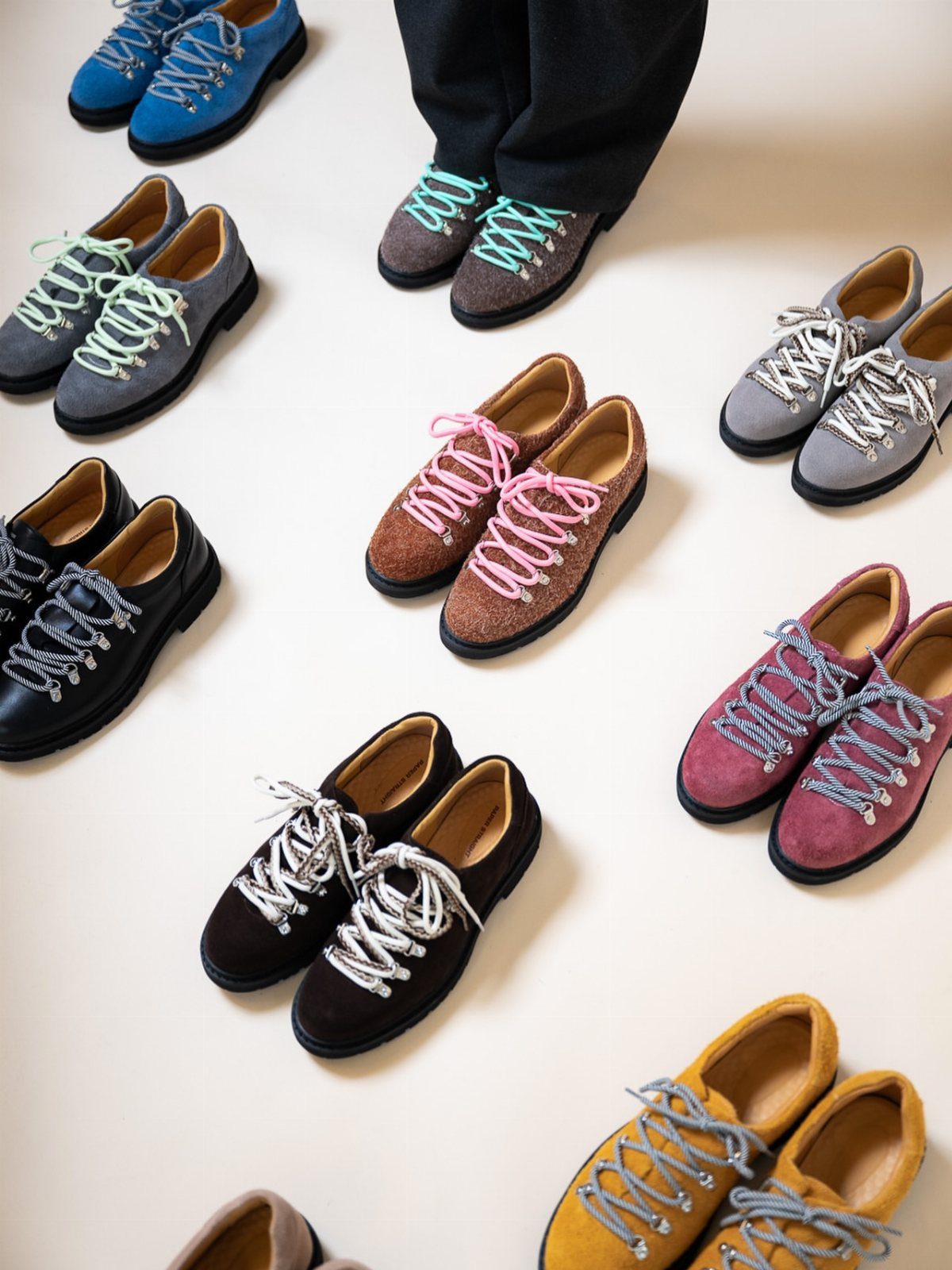 Various colorful suede lace-up shoes arranged on a light neutral background.