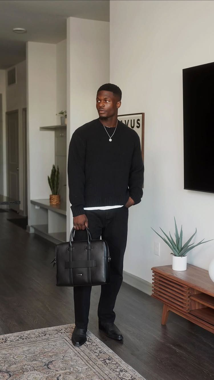 Man wearing a black sweater and dark jeans holding a black leather bag.