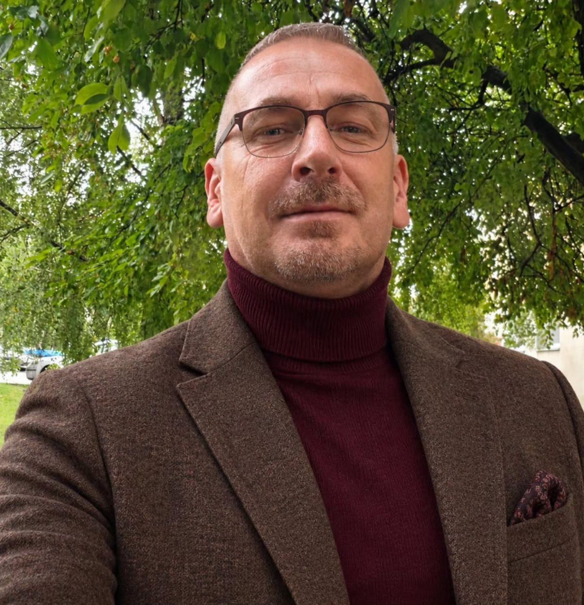 Man wearing a burgundy turtleneck sweater and brown blazer against a green background