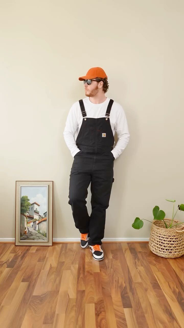 Man wearing black overalls, white shirt, and orange cap leaning against a wall.
