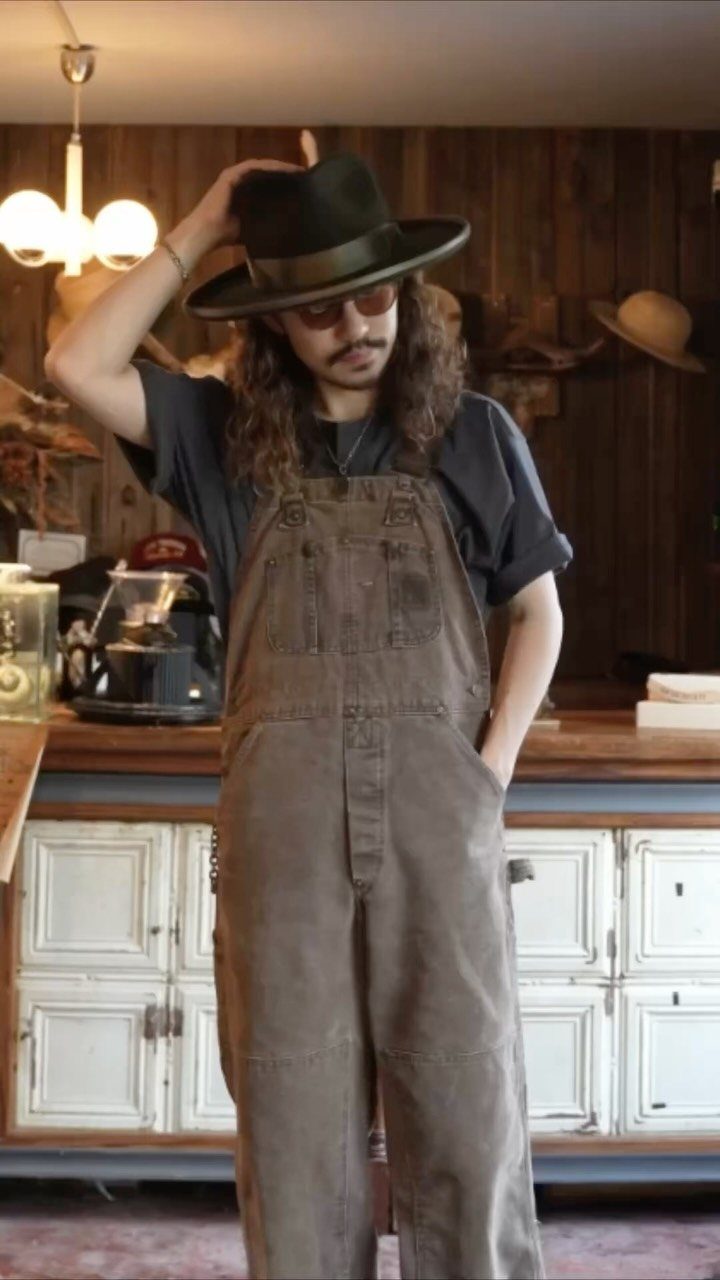 Man wearing brown overalls, a black hat, and a dark t-shirt in rustic interior.