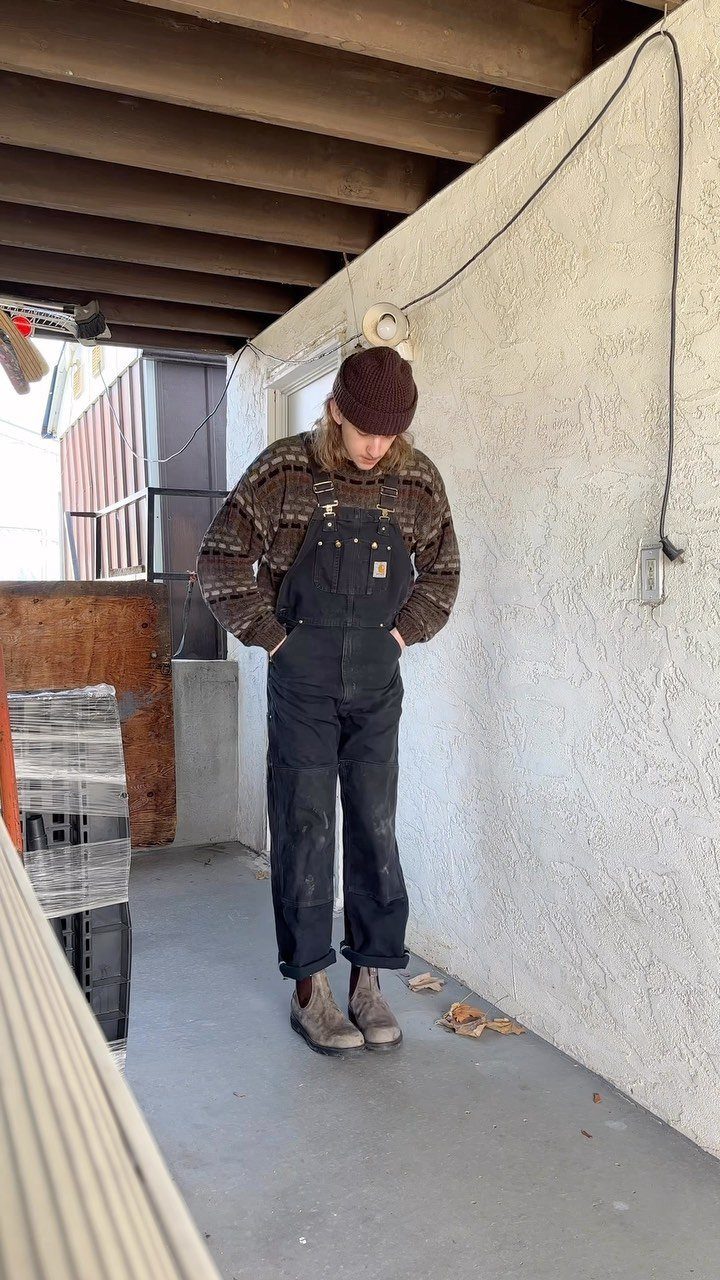 Man wearing dark navy overalls, a patterned brown sweater, and a maroon beanie.