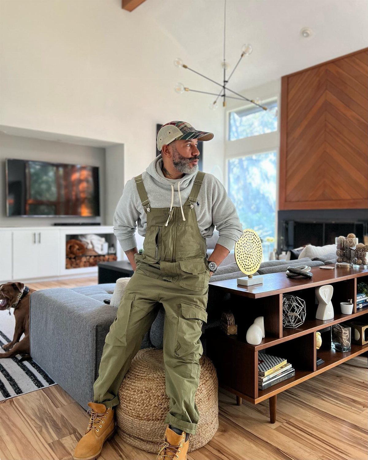 Man wearing olive green overalls, grey hoodie, and tan boots in a living room