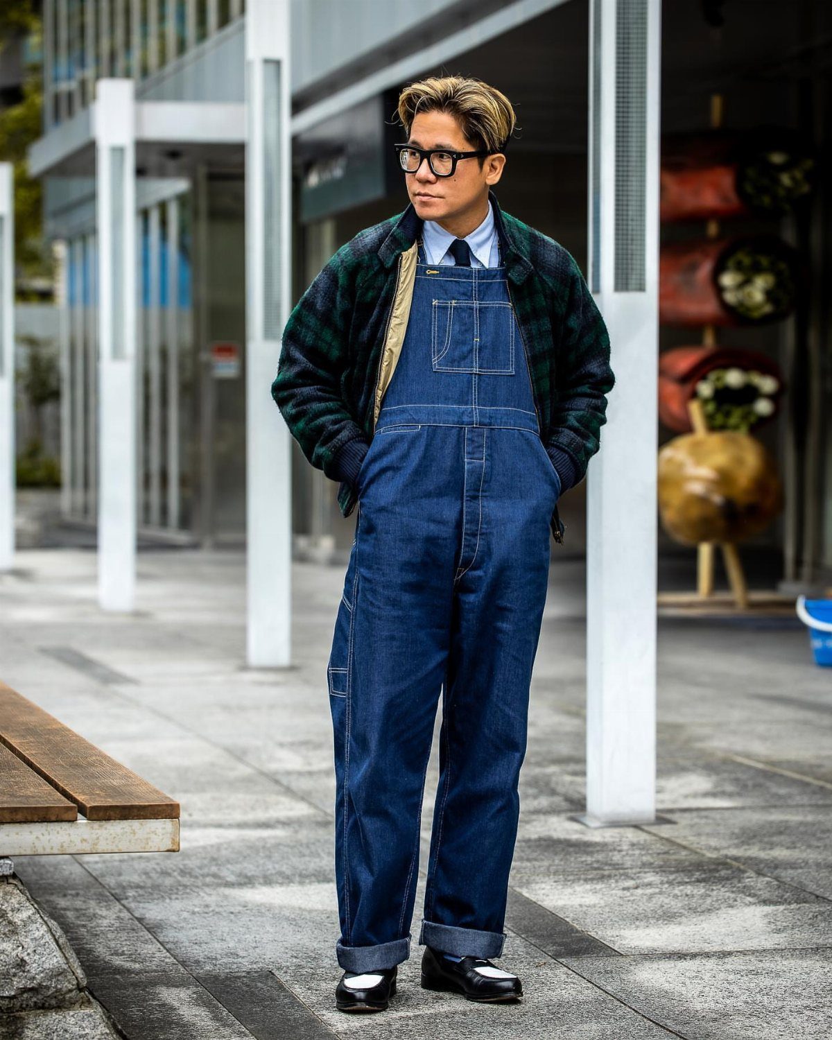 Man in blue denim overalls, plaid jacket, and styled hair standing outdoors.