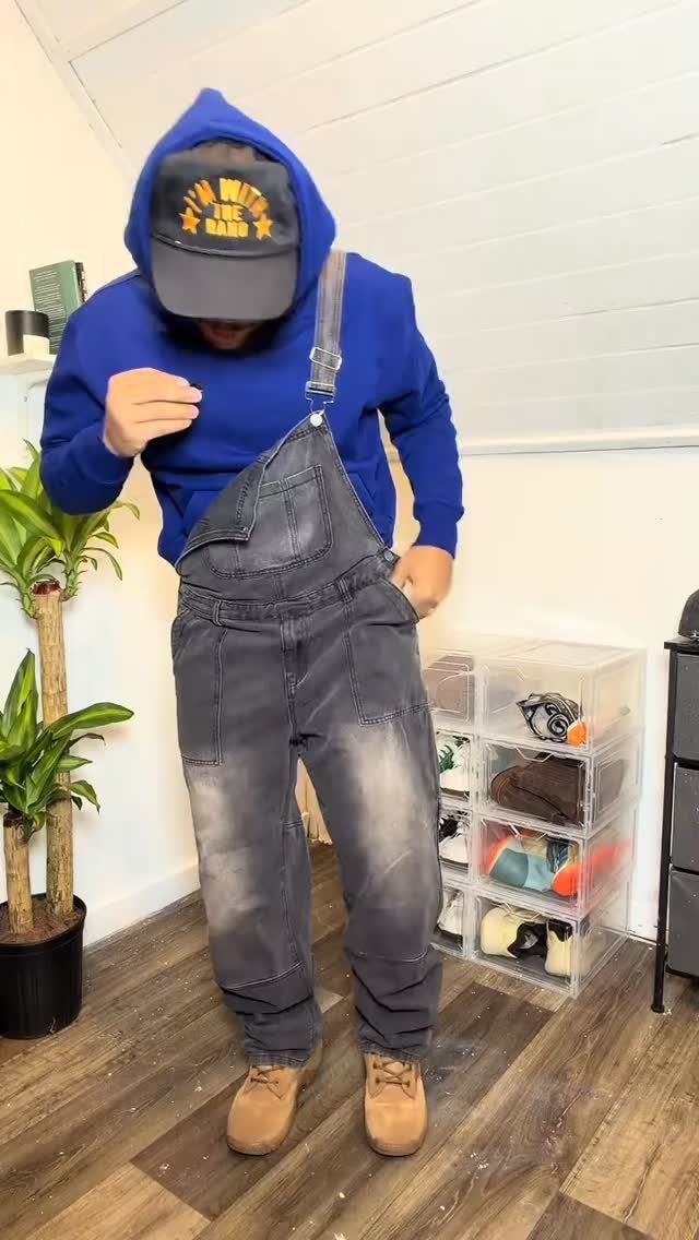 Man wearing grey denim overalls over a blue hoodie and black baseball cap.
