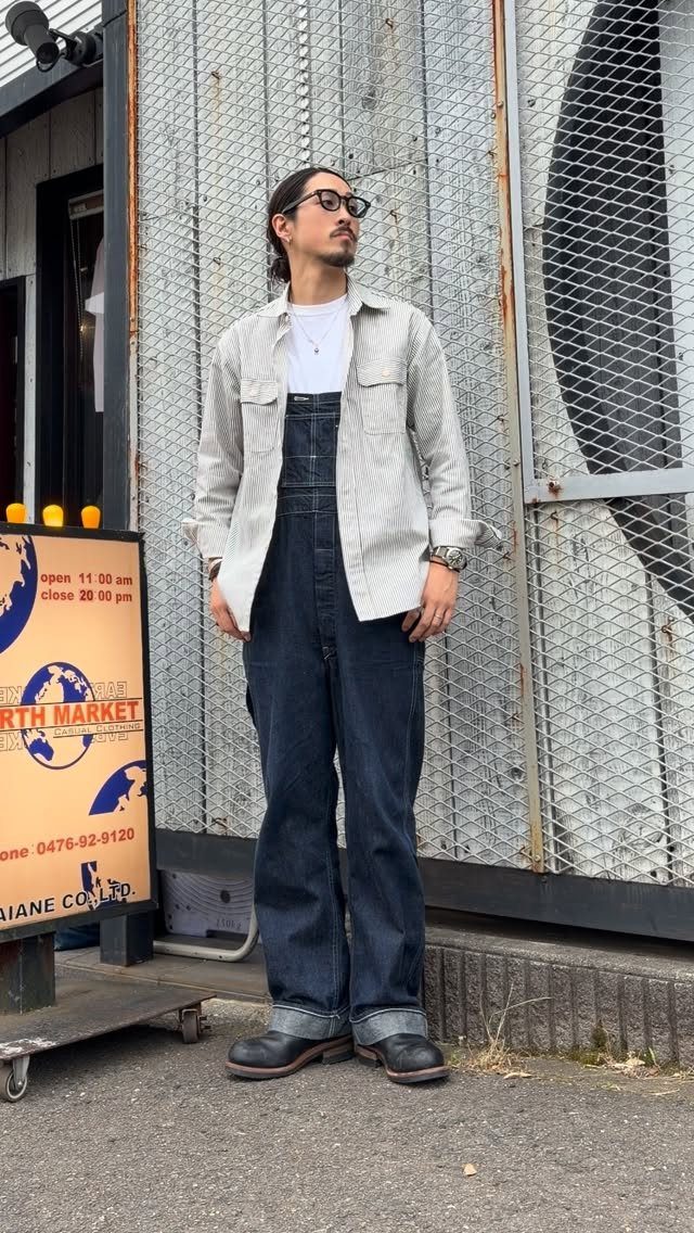 Man wearing dark blue denim overalls with a striped shirt and black shoes.