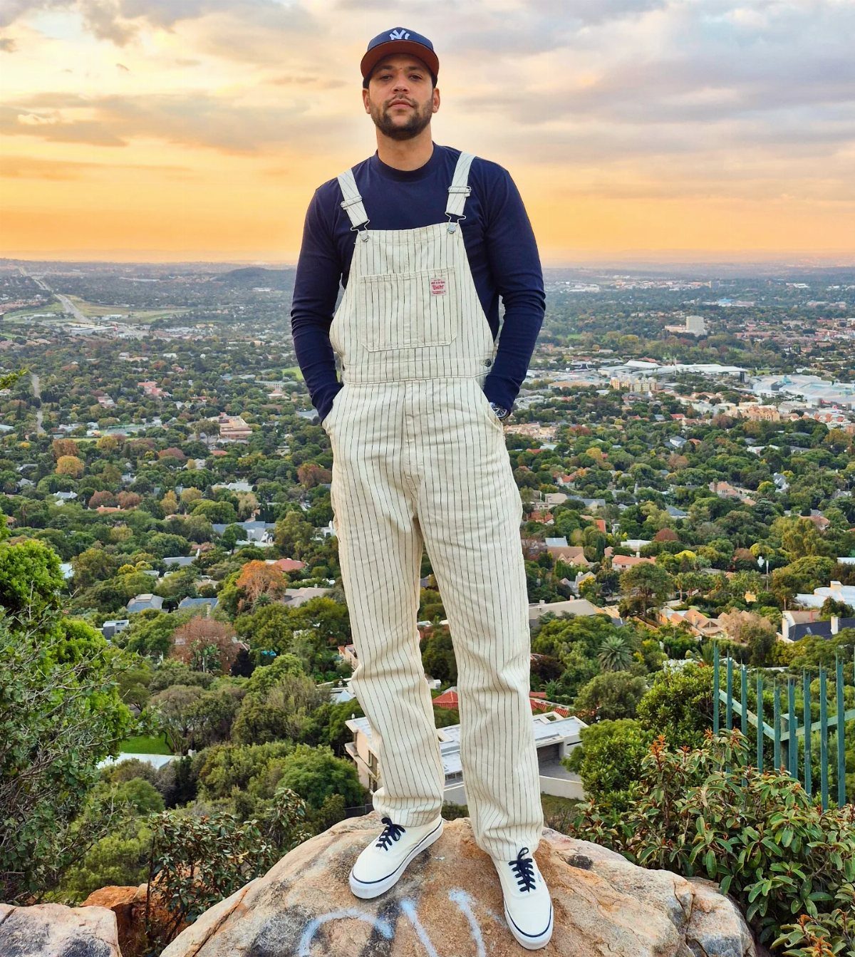 Man in white pinstriped overalls and plain navy shirt standing on a rocky cliff.