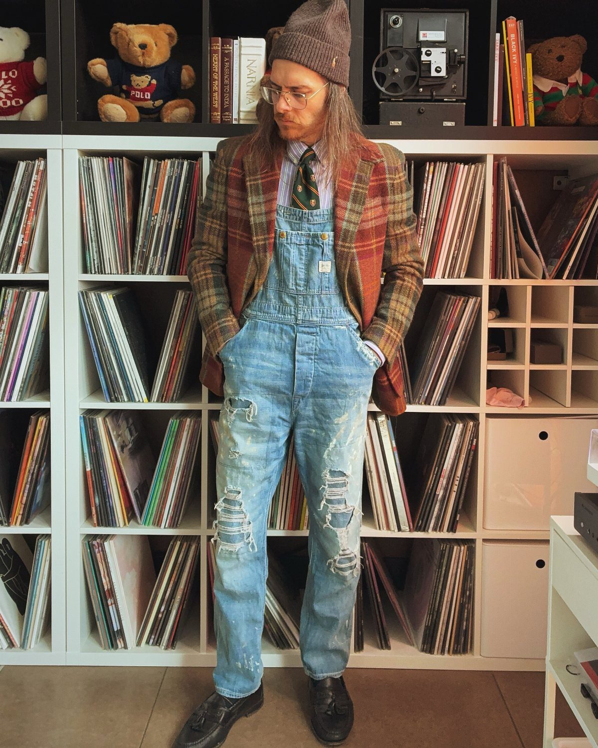 Man in distressed denim overalls, plaid blazer, and beanie standing by record shelves.