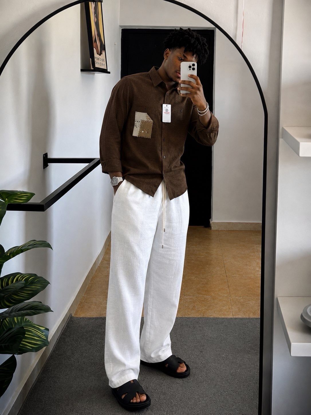 Man in brown button-down shirt and wide white trousers taking a mirror selfie.