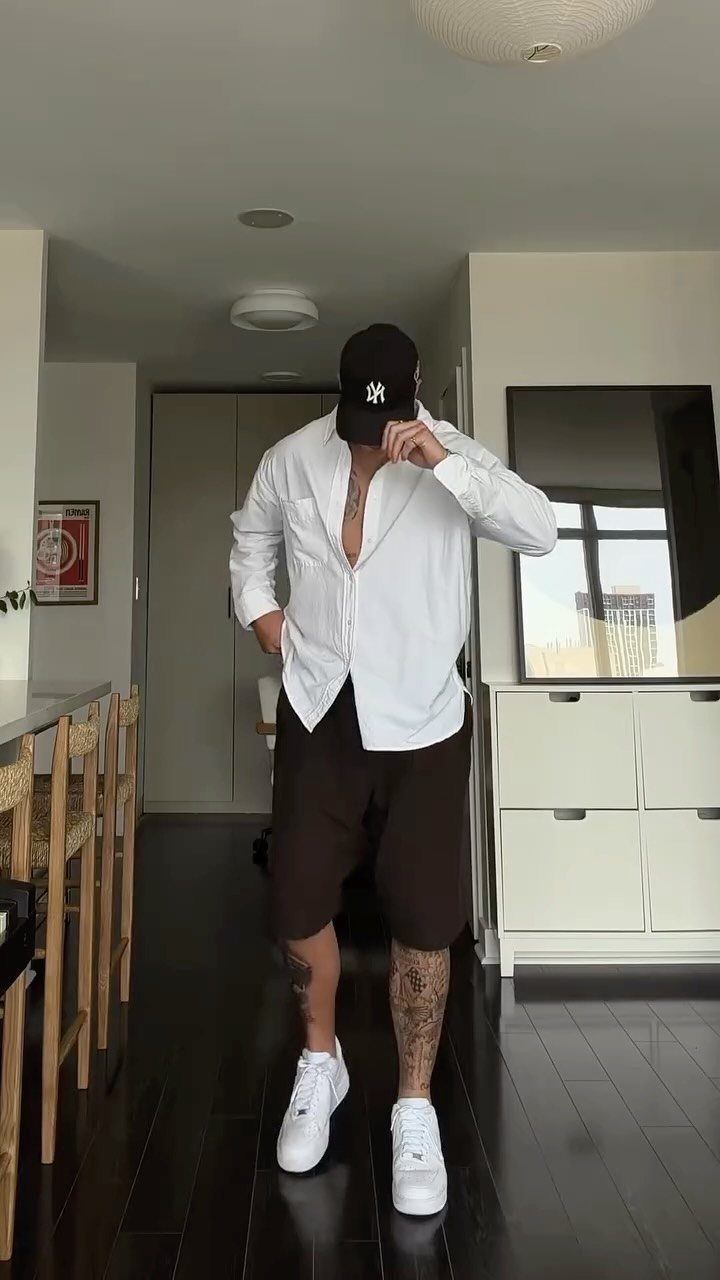 Man wearing a white open shirt, brown shorts, black cap, and white sneakers.