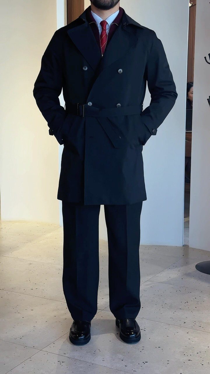 Person in a black buttoned trench coat, black trousers, and a red tie.