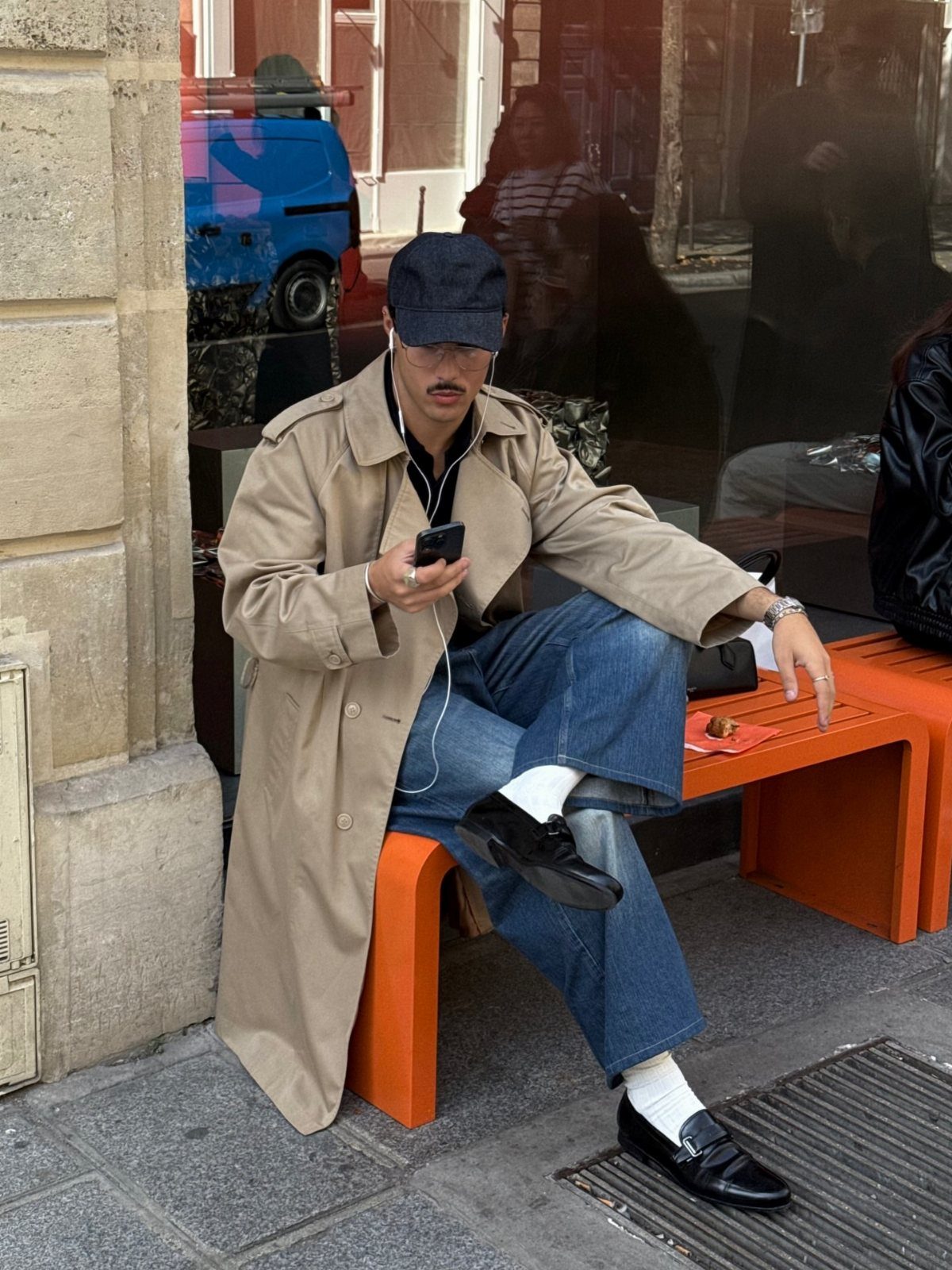 Man in beige trench coat and blue jeans sitting on an orange bench.