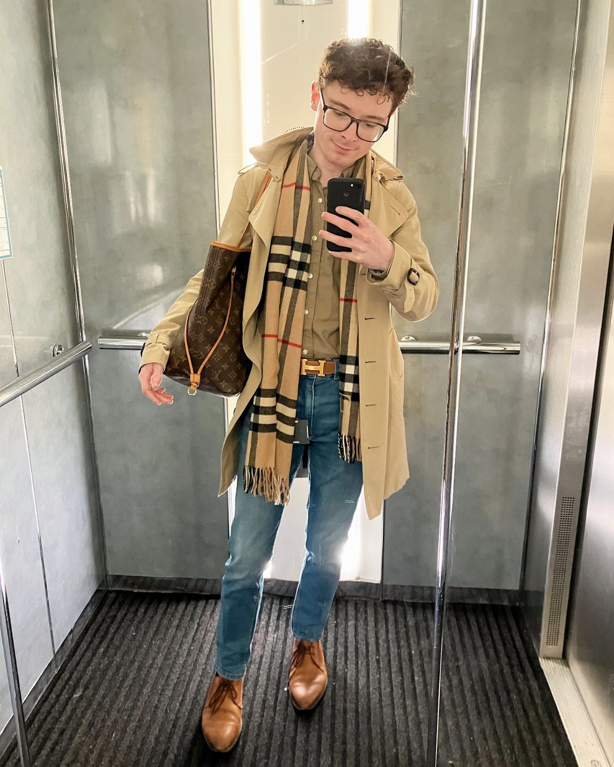 Person in a beige trench coat and plaid scarf posing in an elevator mirror.
