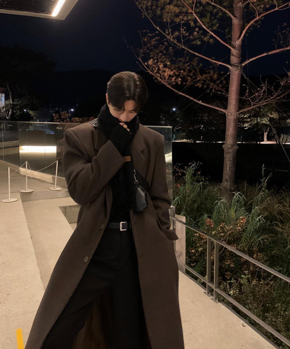Man in a long brown trench coat and black turtleneck standing outdoors at night.