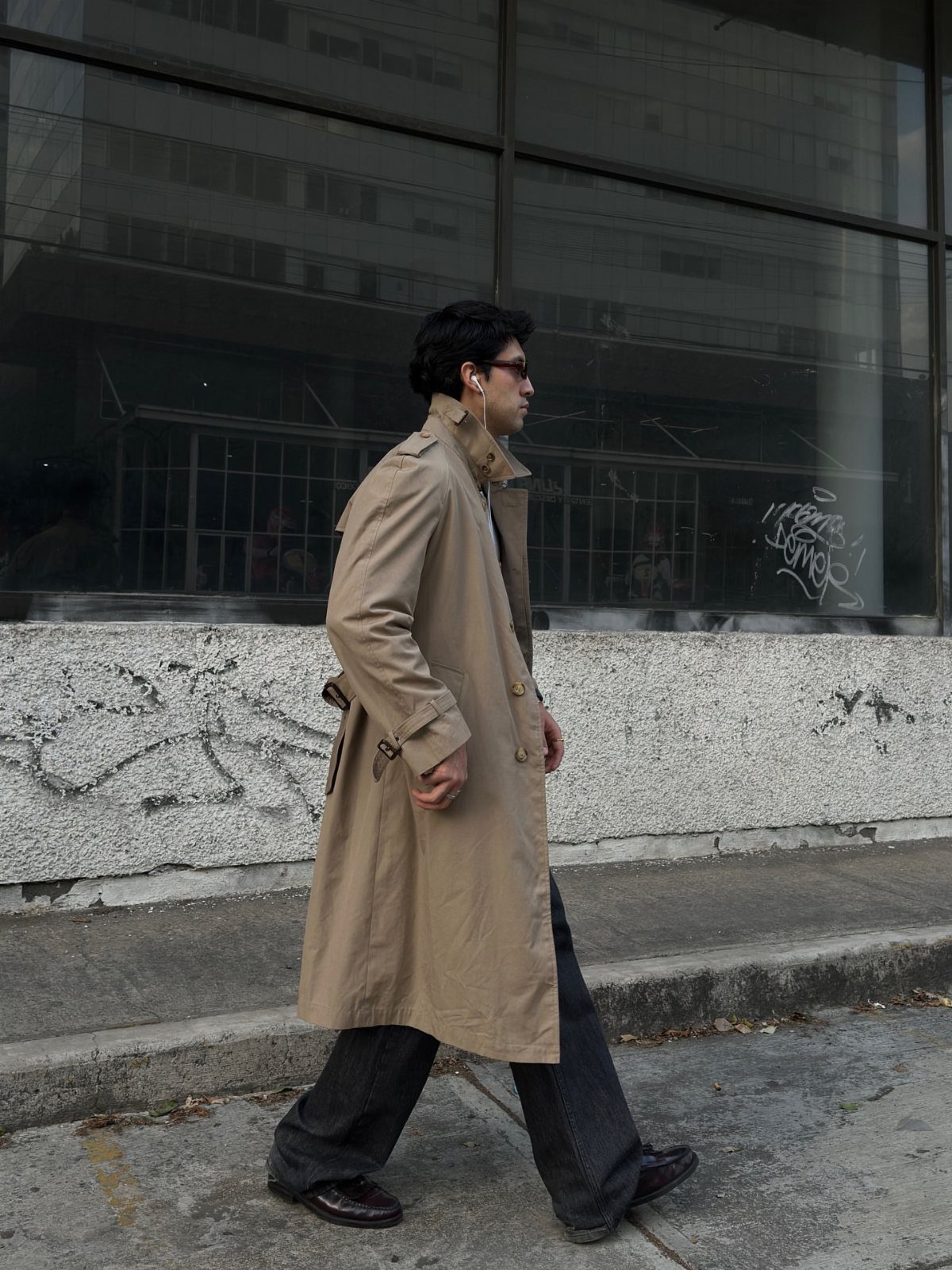 Man in a tan trench coat and wide-leg trousers walking on a city street.