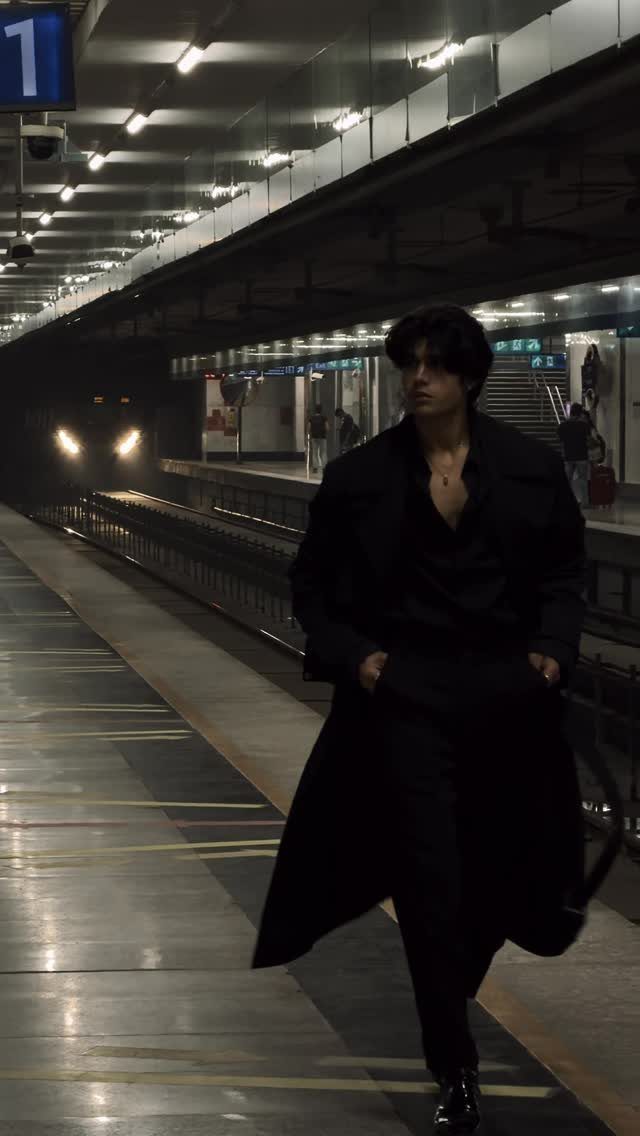Man in a long black trench coat standing on a train station platform.