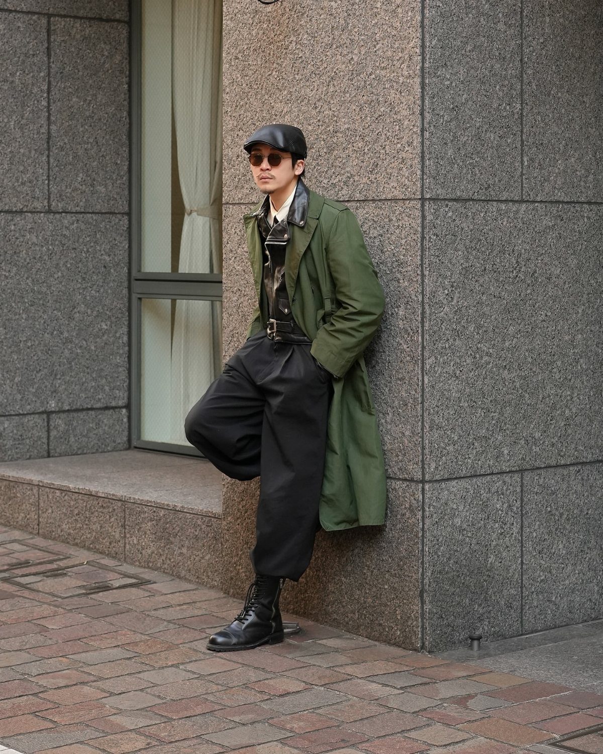 Man in olive green trench coat and black combat boots leaning against stone wall