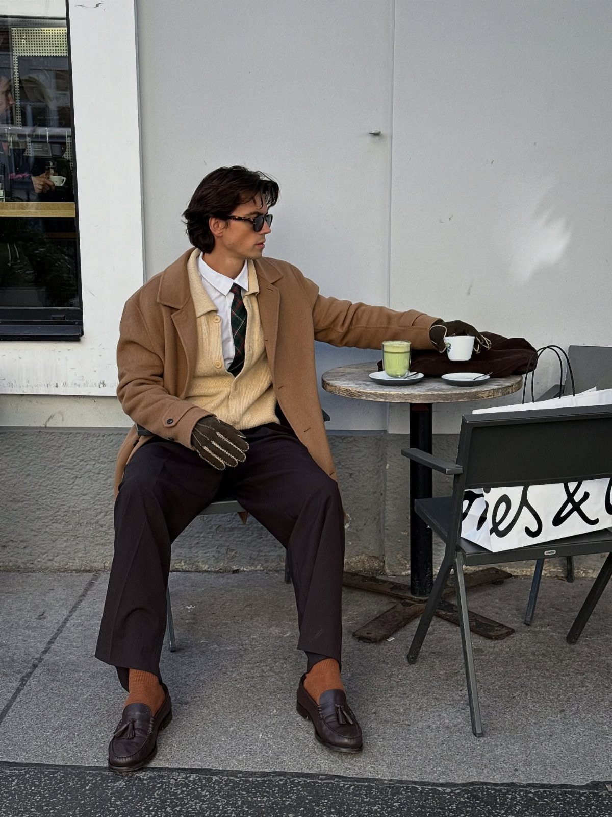 Man in tan trench coat and cream cardigan sitting at outdoor cafe table
