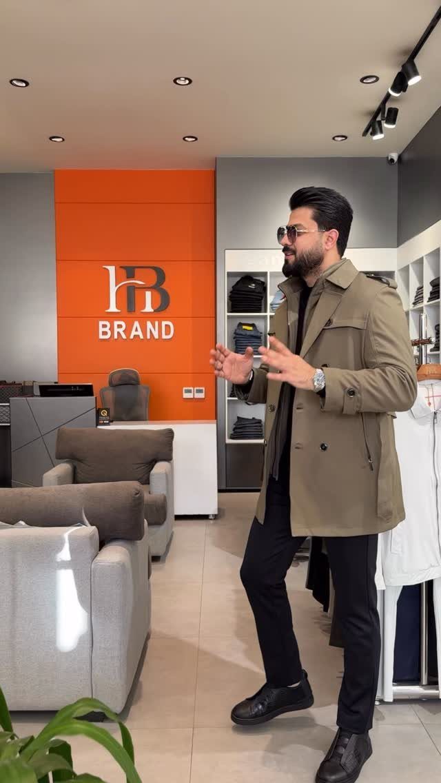 Man in a tan trench coat and black trousers standing in a clothing store.