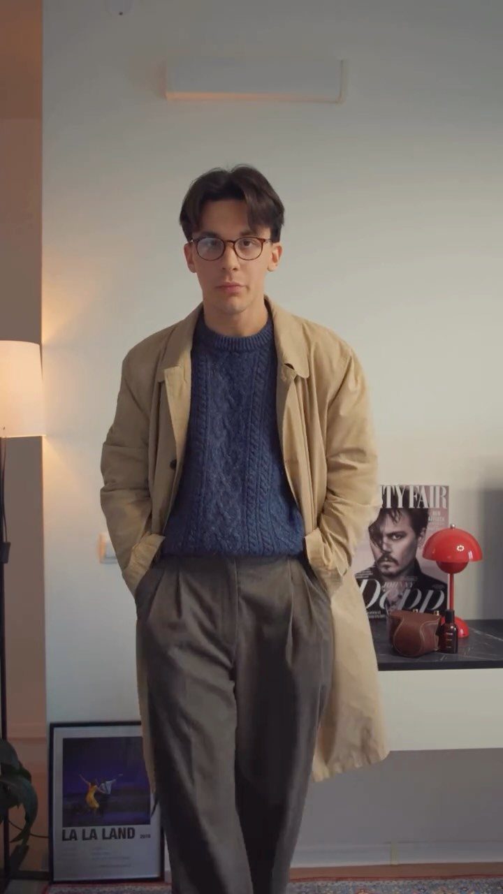 Man wearing a tan trench coat over a blue cable-knit sweater and grey trousers.