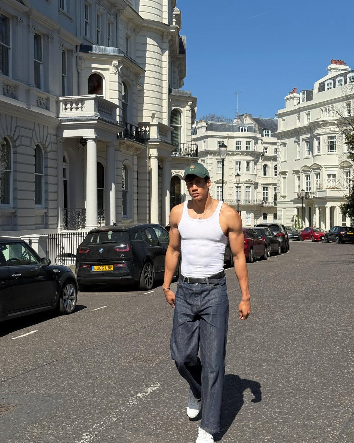 Muscular man in white tank top and blue trousers walking on city street